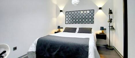 Un dormitorio moderno con tragaluz, cama con colcha blanco y negro, dos mesitas de noche con lámparas y una lámpara de pared.
