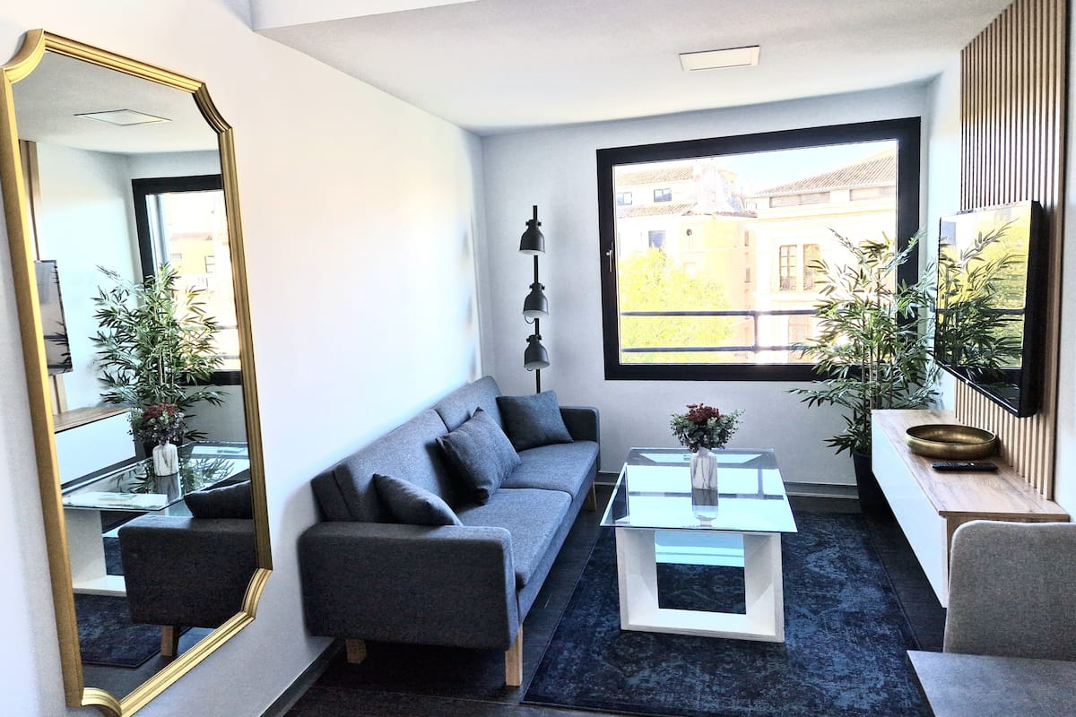 Ein modernes Wohnzimmer mit einem grauen Sofa, einem Glastisch und einem großen Fenster mit Blick auf Gebäude.