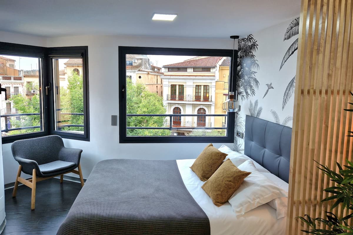 Ein Schlafzimmer mit einem großen Bett, zwei Kissen, einem Sessel, einem Fenster mit Blick auf Gebäude und einer Deckenleuchte.