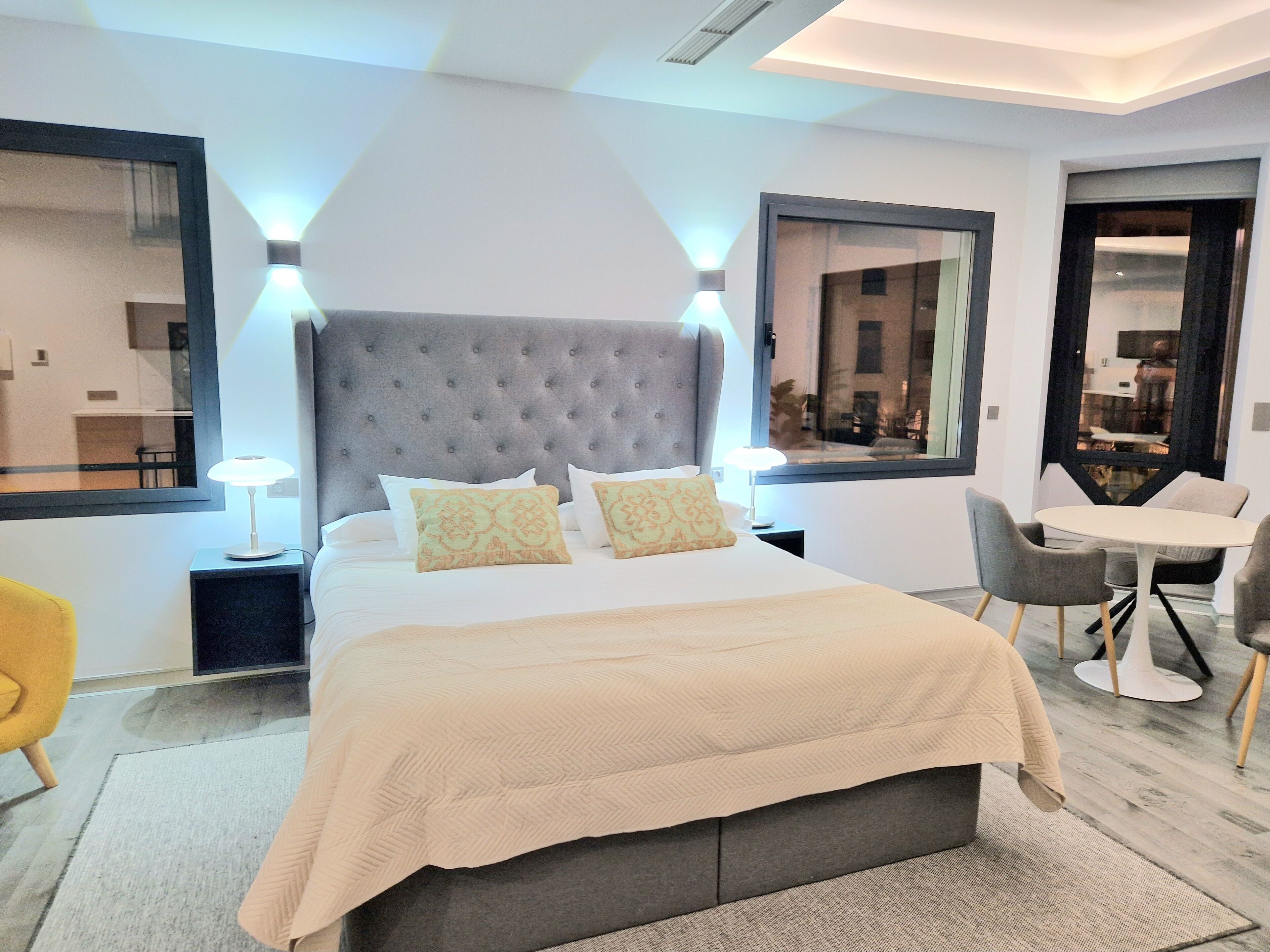 Una habitación de hotel moderna con una cama grande, dos lámparas de noche, un cabecero tapizado en gris y una pequeña mesa con sillas.