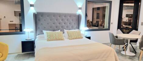 Una habitación de hotel moderna con una cama grande, dos lámparas de noche, un cabecero tapizado en gris y una pequeña mesa con sillas.