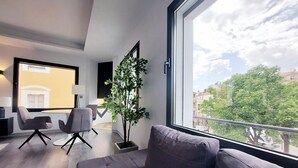 Een moderne woonkamer met een grijze bank, een kleine tafel en twee stoelen. Een groot raam biedt uitzicht op gebouwen en bomen.