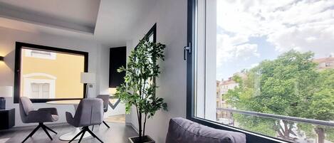 Una sala de estar moderna con un sofá gris, una mesita y dos sillones. Un ventanal ofrece vistas a edificios y árboles.