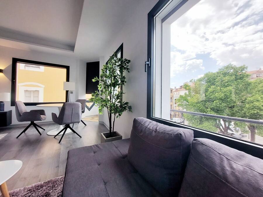 Una sala de estar moderna con un sofá gris, una mesita y dos sillones. Un ventanal ofrece vistas a edificios y árboles.