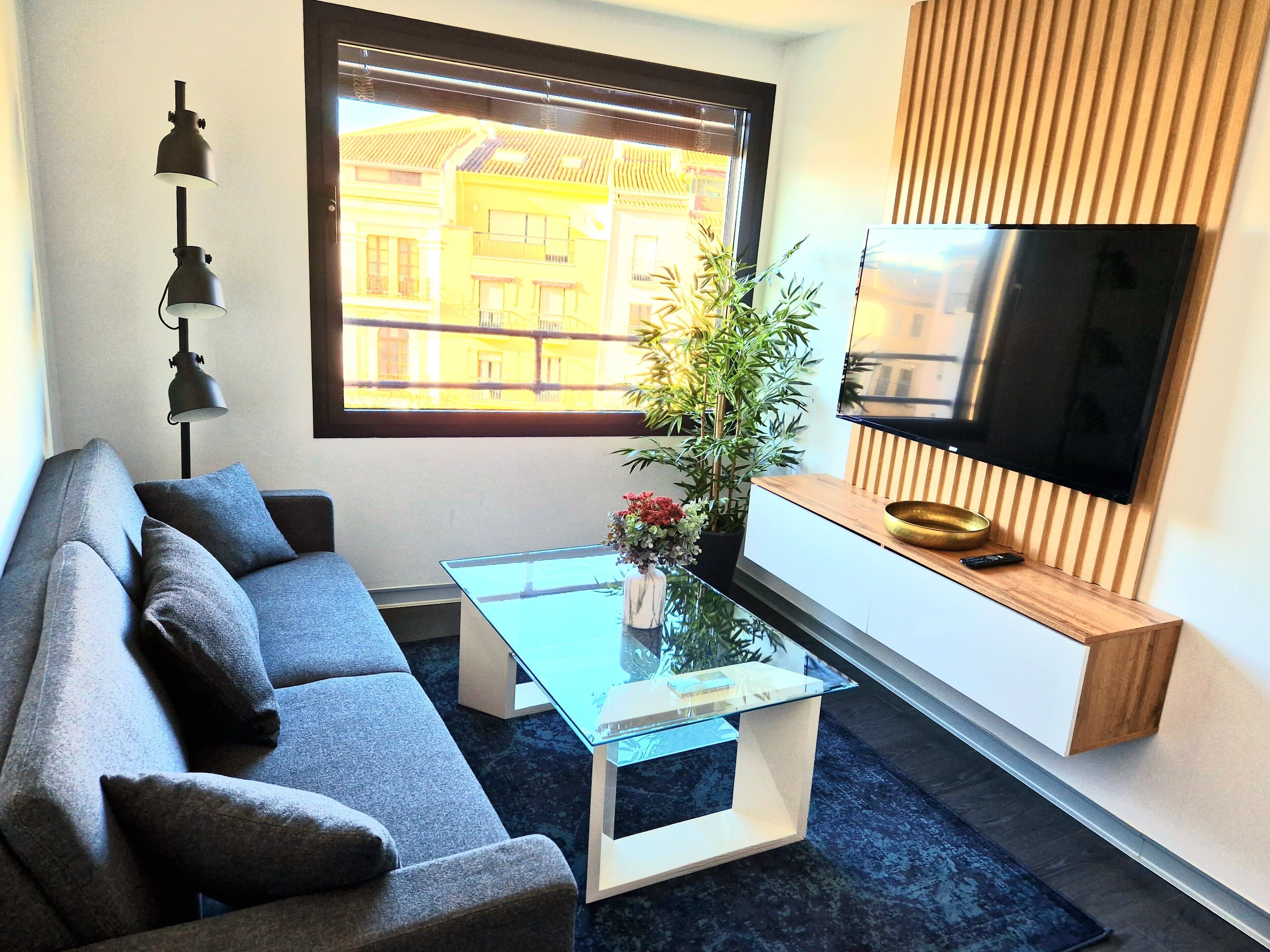 Una sala de estar moderna con un sofá, una mesa de centro de vidrio, una planta en maceta y un televisor de pantalla plana montado en la pared.