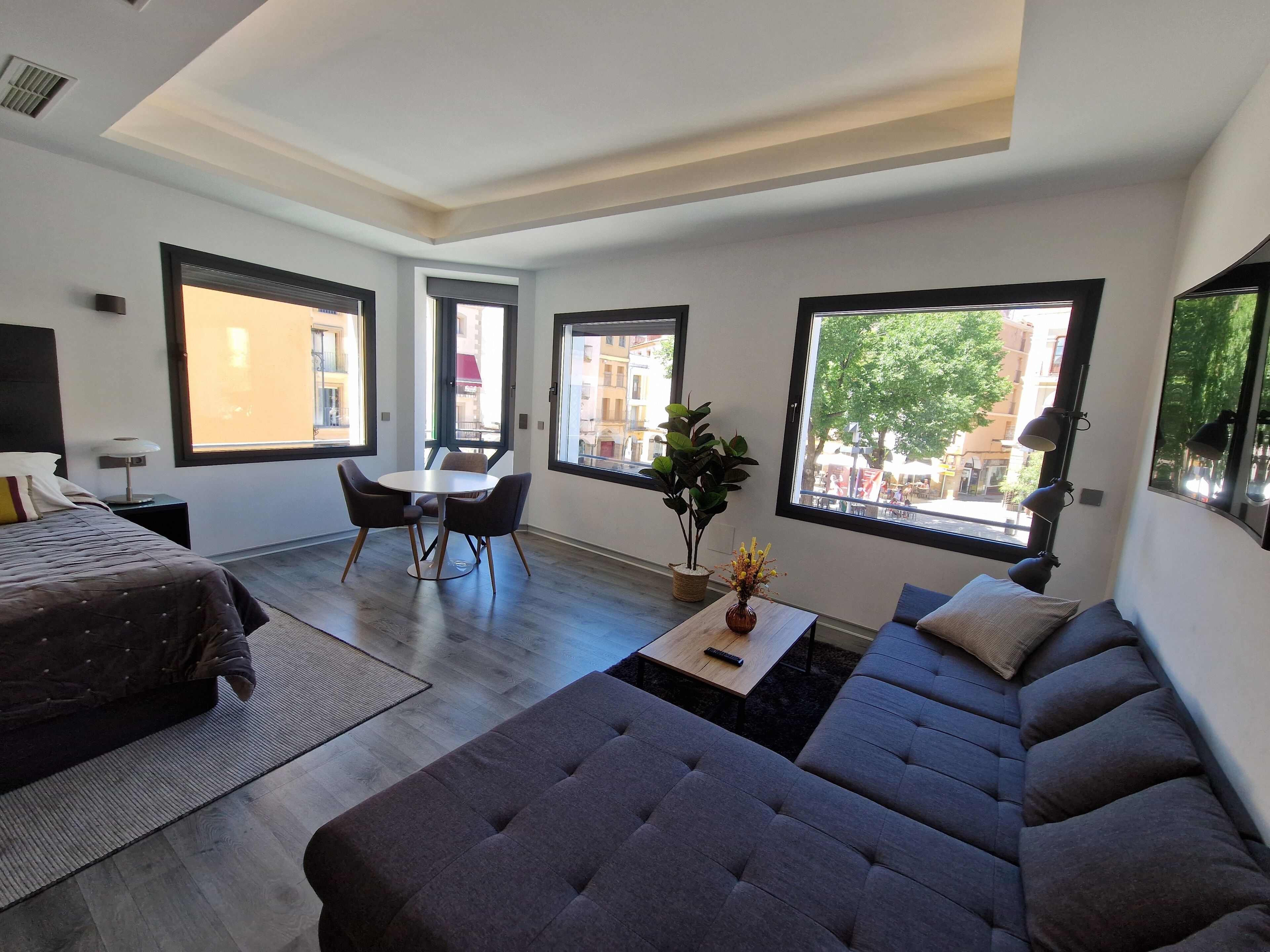 A modern living room with a sofa, a dining table, and chairs. There are large windows providing views of the city.