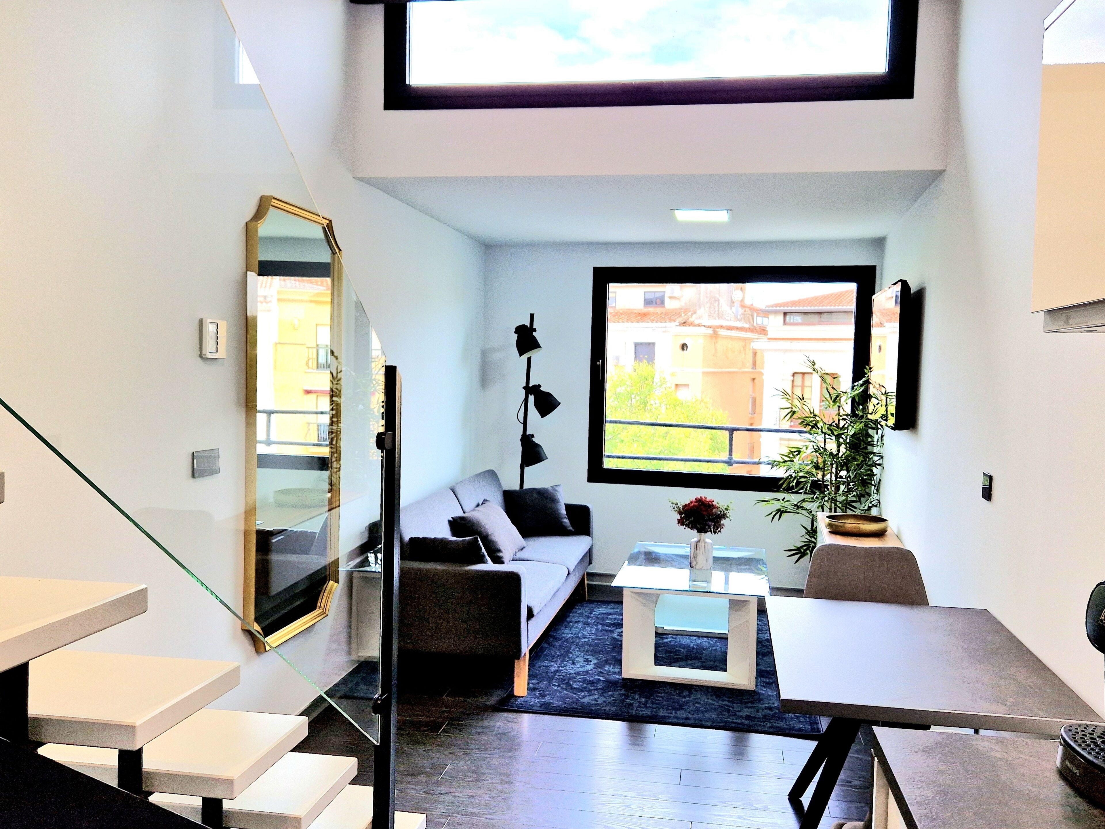 A modern living room with a sofa, coffee table, and a potted plant. There is a staircase with a glass railing leading to an upper level.