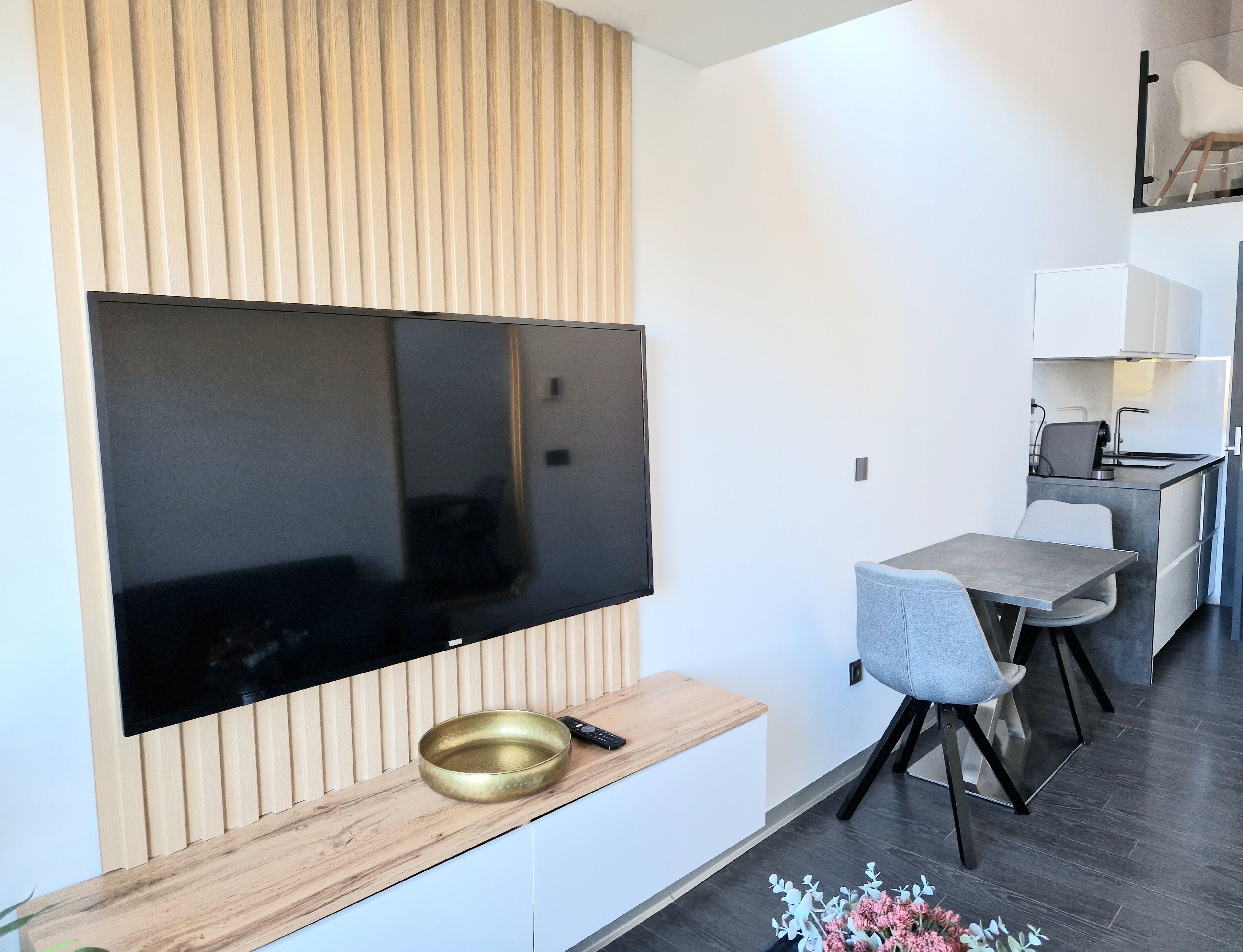 A modern living space with a large flat-screen TV mounted on a wooden panel wall, a small wooden table with a bowl and remote, a grey upholstered chair, and a compact kitchenette in the background.