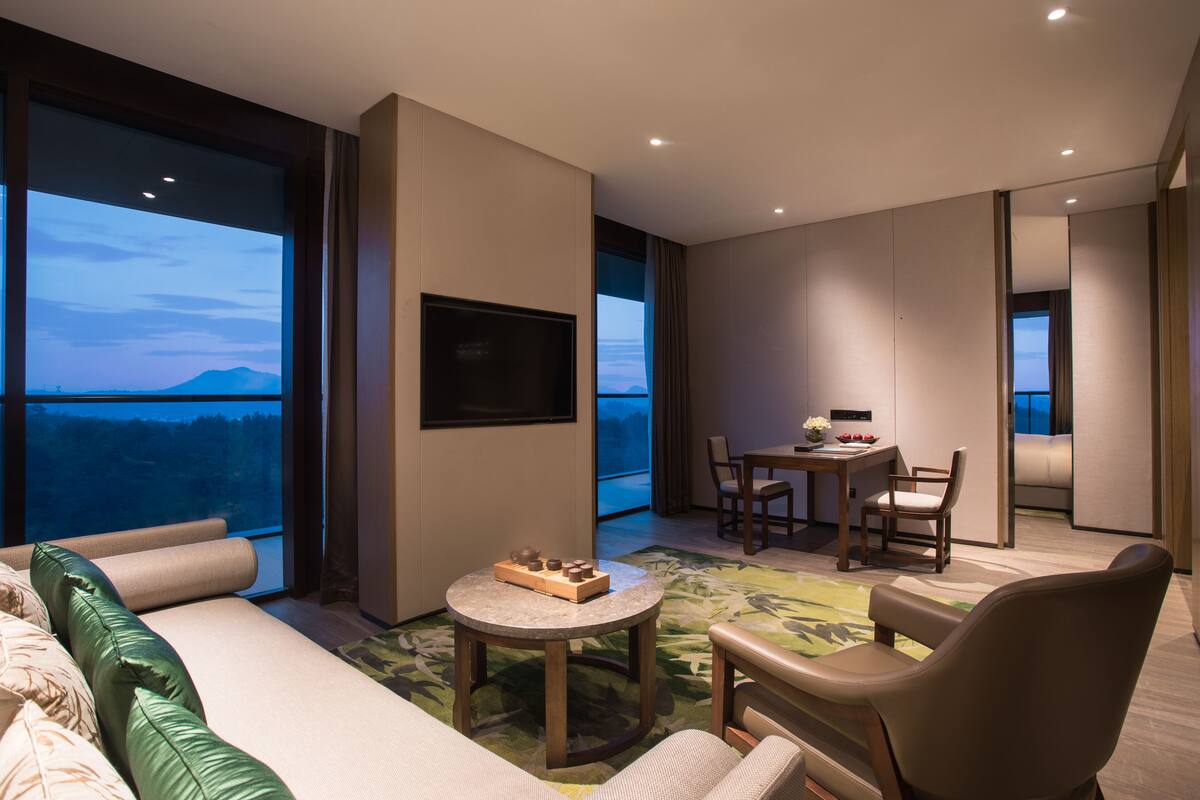 A modern hotel room with a large window offering a mountain view, a flat-screen TV mounted on the wall, a dining area with a table and chairs, and a cozy seating area with a sofa and armchairs.