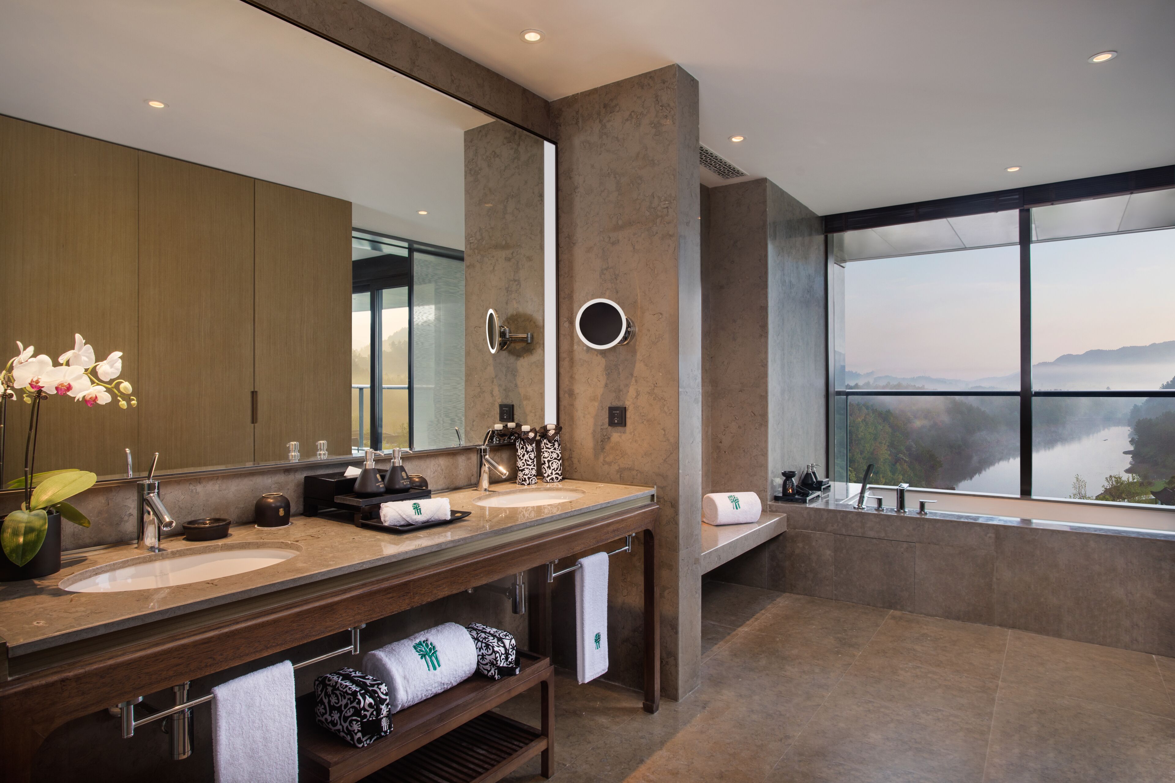 A modern bathroom with dual sinks, a large mirror, and a bathtub with a view of a lake.