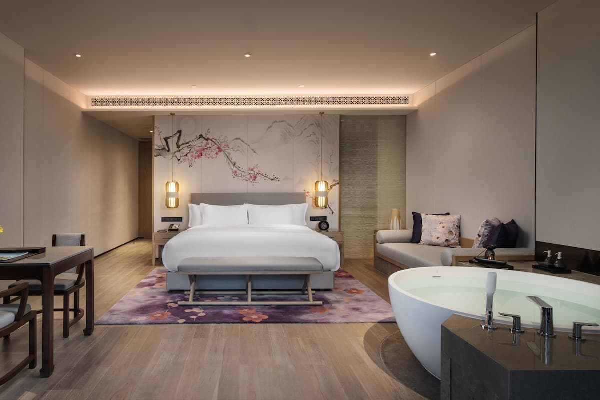A modern hotel room with a large bed, a desk, a chair, a sofa, and a bathtub.
