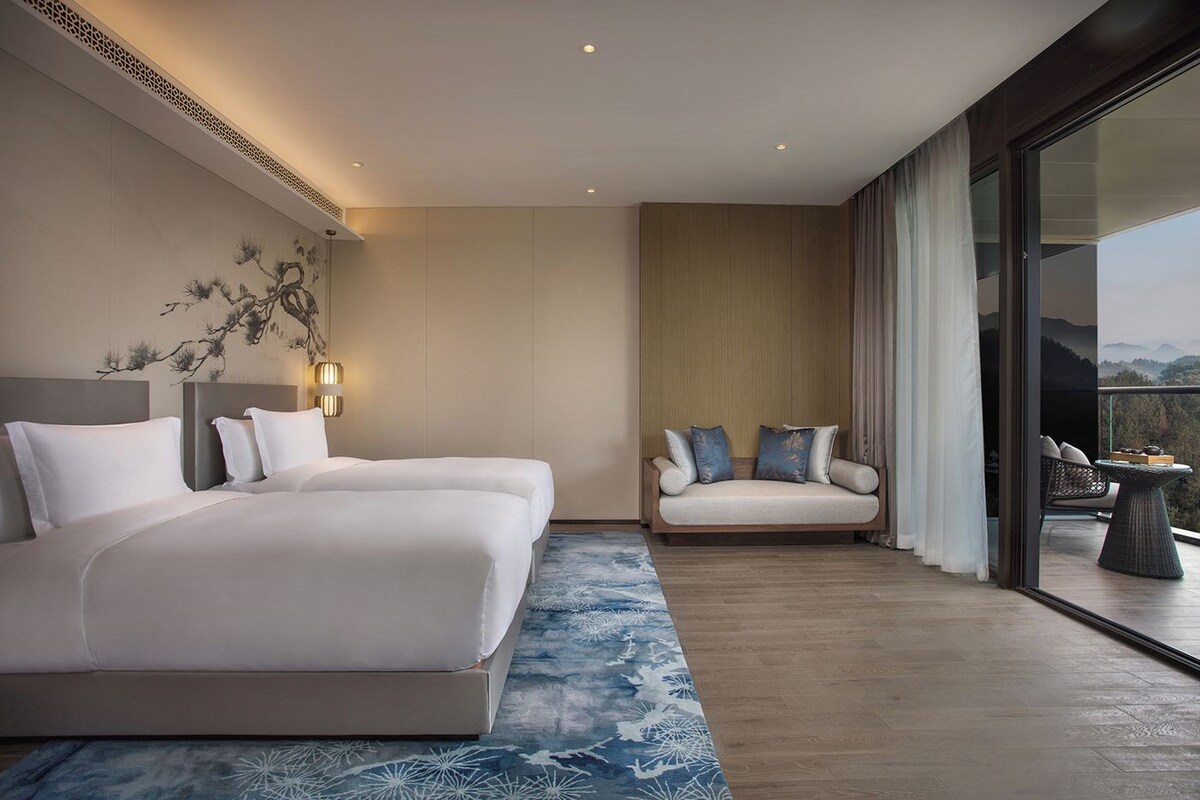 Habitación de hotel con una cama grande, un sofá, un balcón con vistas y un elemento decorativo en la pared.