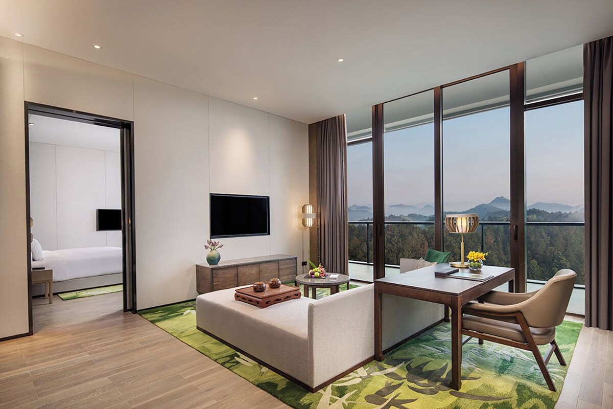 Una habitación de hotel moderna con una cama, un televisor, un área de comedor con una mesa y sillas, y un ventanal con vistas al exterior.