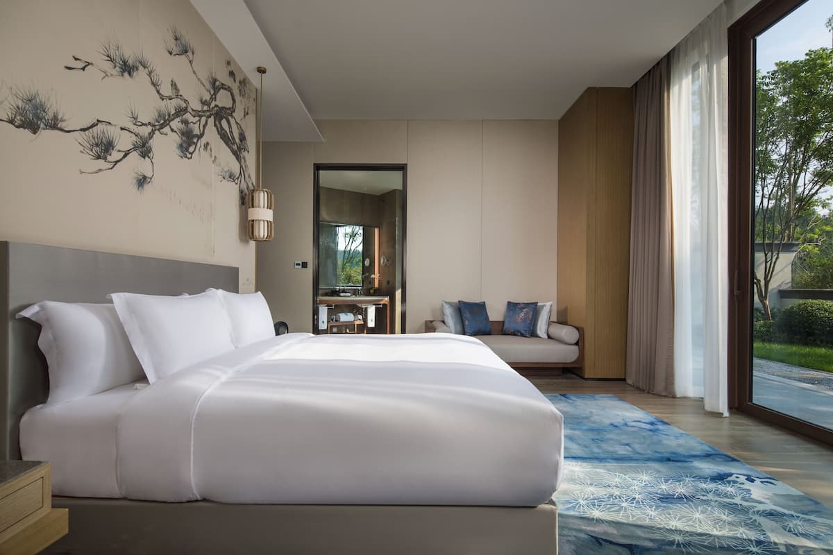 A modern hotel room with a large bed, a sofa, a dining area, and a view of the outdoors.