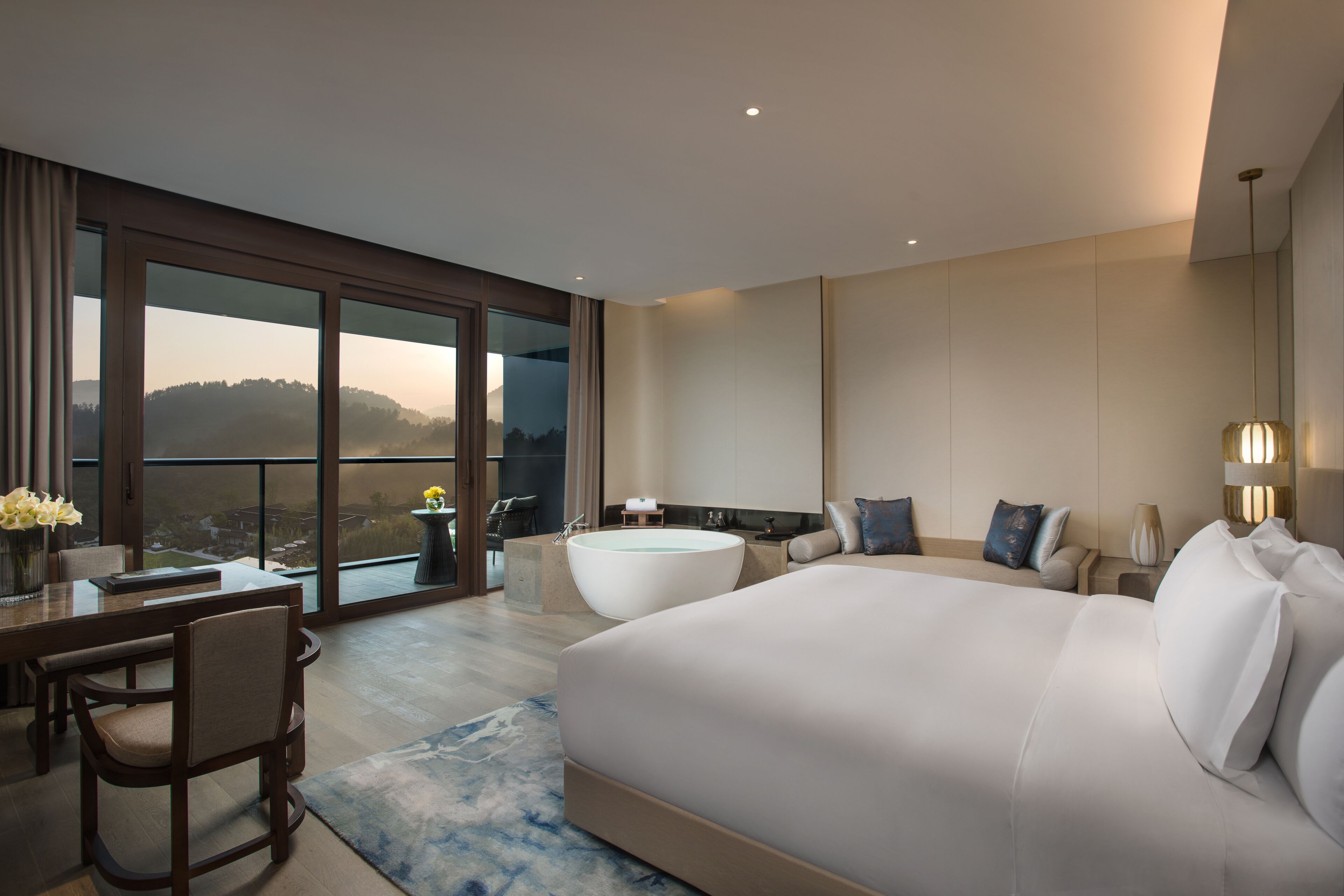 A modern hotel room with a large bed, a sitting area with a sofa and armchairs, a dining table with chairs, a large window with a view of the hills, and a bathtub.