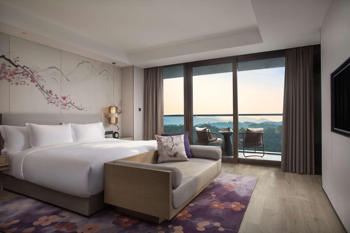 Habitación de hotel con una cama grande, un sofá, una mesita y vistas a la montaña a través del ventanal.