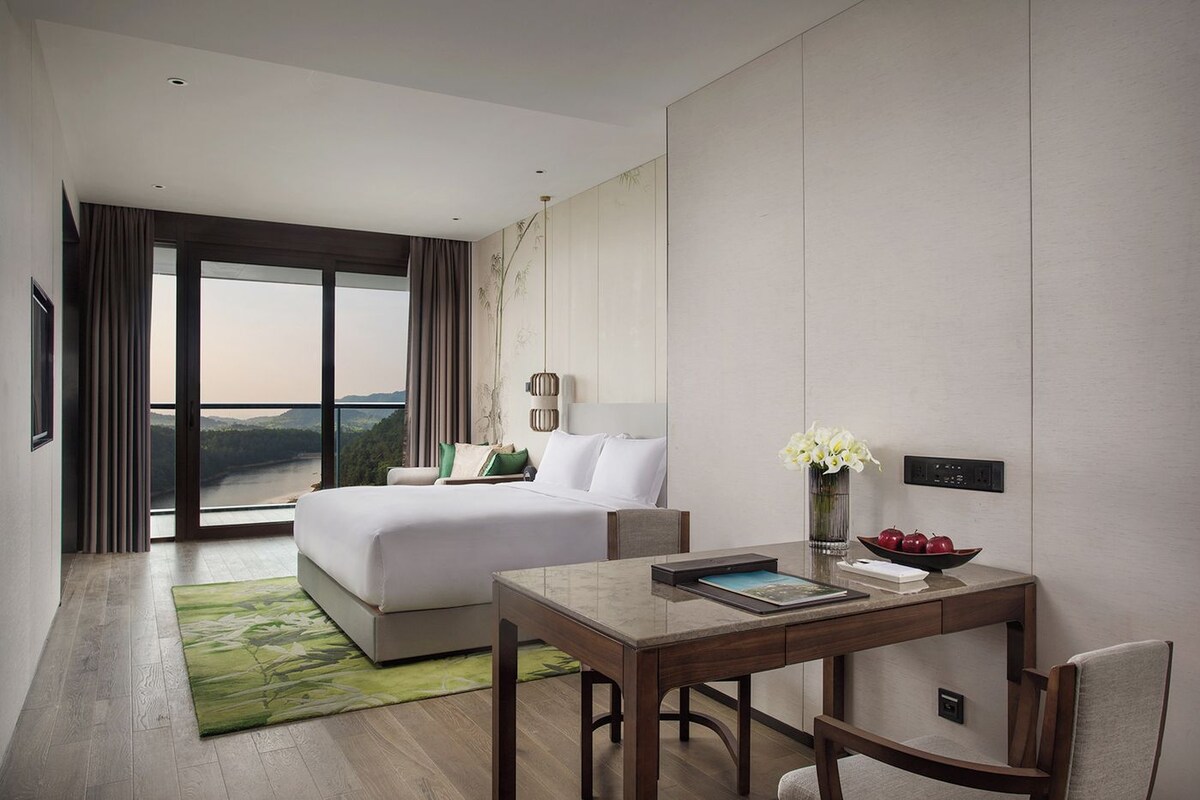 Una habitación de hotel moderna con una cama grande, una mesa de comedor y vistas a un paisaje natural.