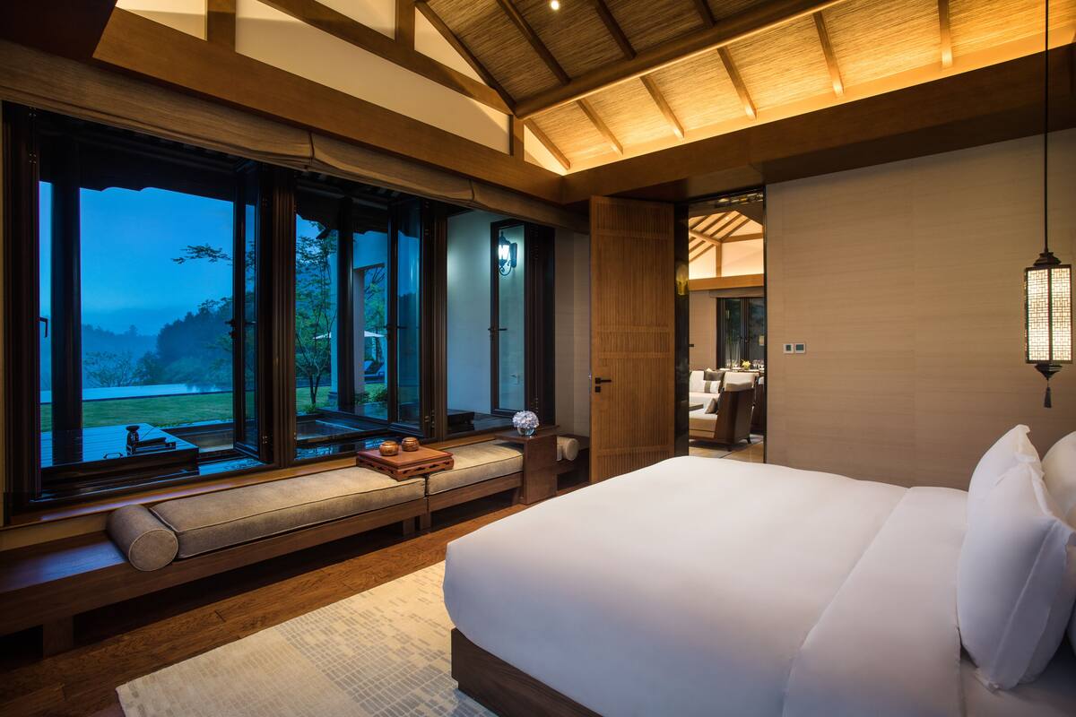 A bedroom with a large bed, a bench with cushions, and a view of a garden through large windows.