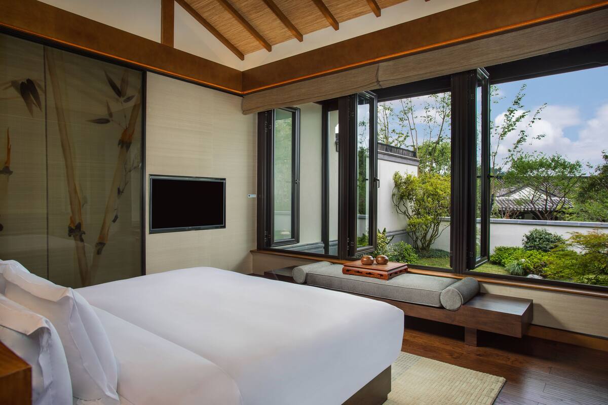 A modern bedroom with a large bed, a TV, and a view of greenery outside.
