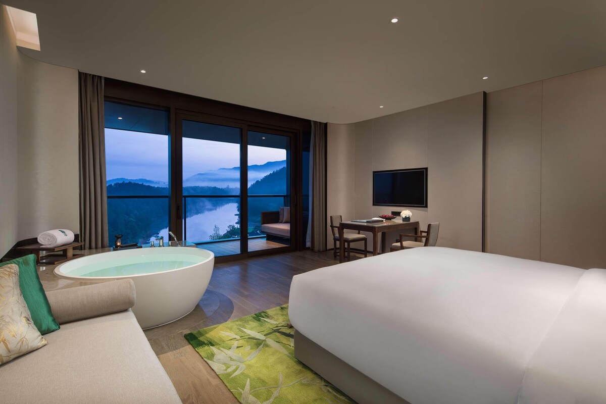 Habitación de hotel moderna con una cama grande, una zona de estar con vistas, una bañera y una mesa de comedor.