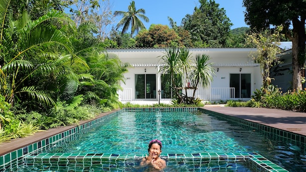 Une piscine à l’eau d’un bleu cristallin, entourée d’une végétation luxuriante et bordée d’un bâtiment blanc au toit recouvert de tuiles.