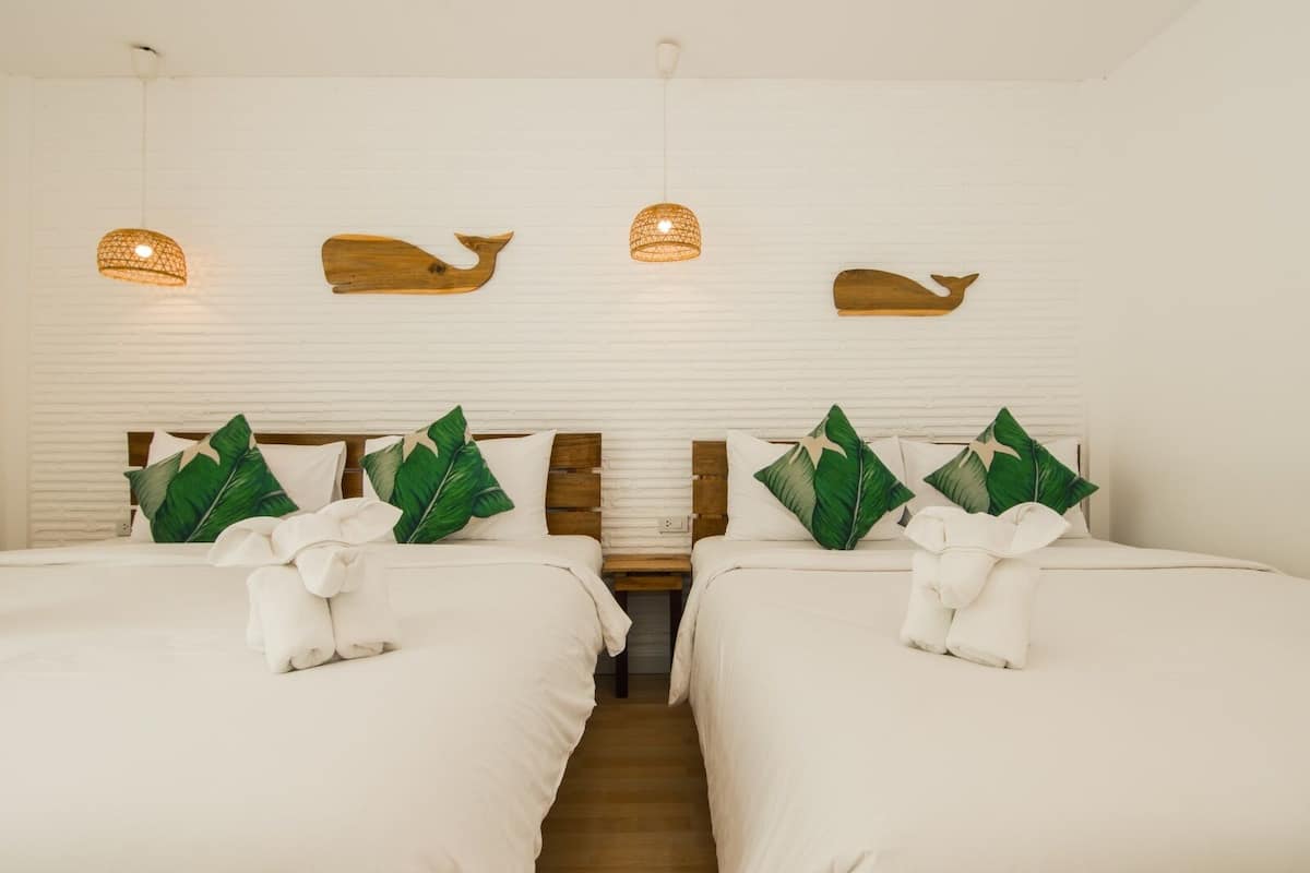 Quarto de hotel com duas camas, roupa de cama branca, almofadas com folhas verdes, cabeceiras de madeira e luminárias pendentes.