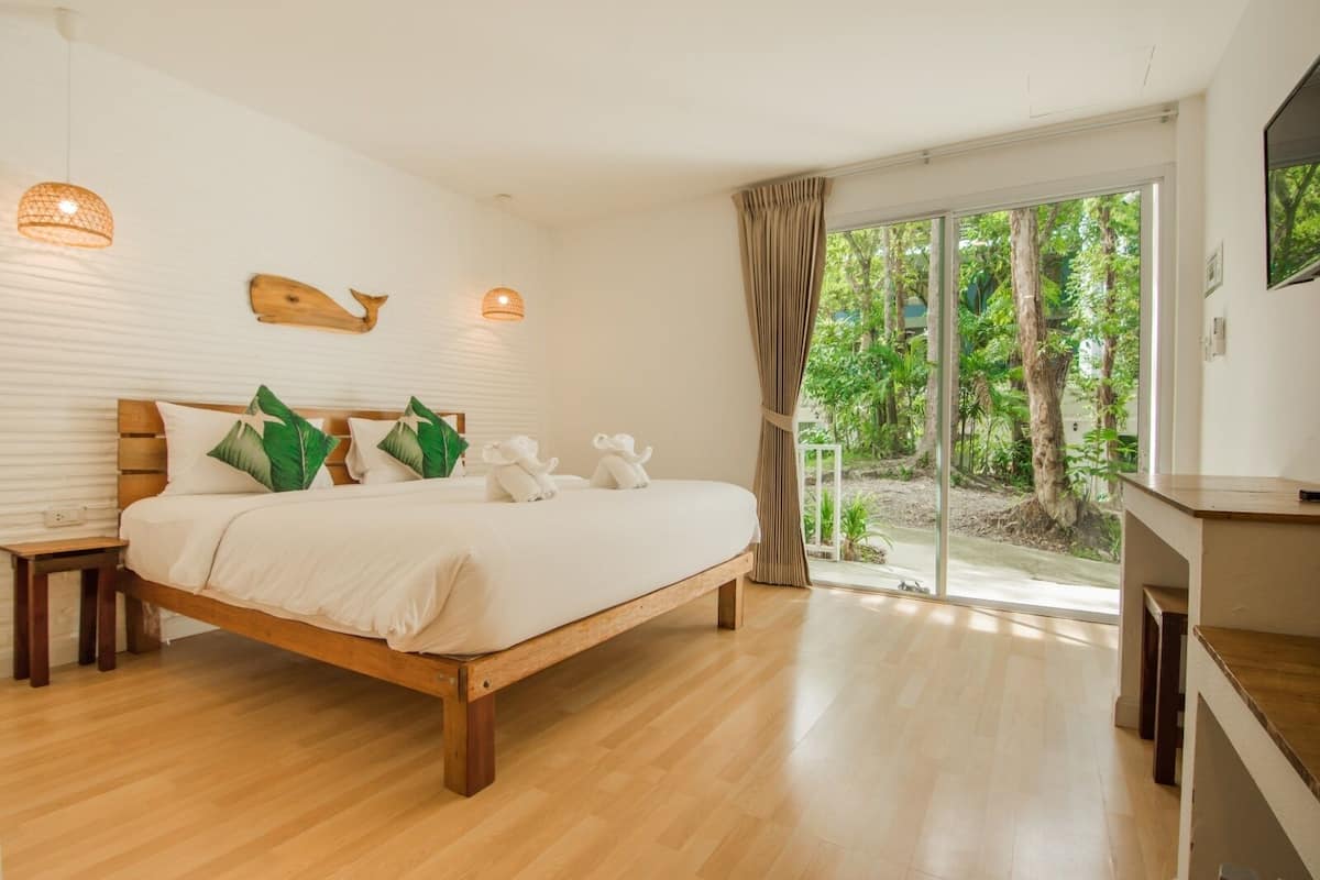 Um quarto espaçoso com uma cama grande, cabeceira de madeira e vista para um jardim exuberante através de uma porta de vidro deslizante.