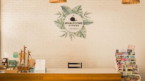 Un comptoir d’accueil au Whalecome Aonang Resort, avec un mur en briques blanches, une pancarte de bienvenue, un banc en bois et un présentoir à magazines.