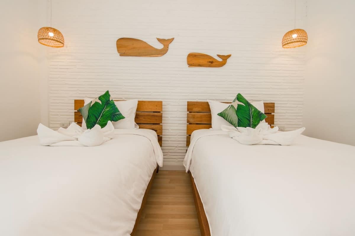 Quarto de hotel com duas camas, roupa de cama branca, cabeceiras de madeira e duas luminárias pendentes fixadas na parede.