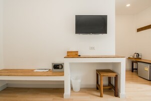 Een kleine, schone kamer met een houten tafel, een witte bank, een aan de muur bevestigde televisie en een kitchenette met een koelkast en koffiezetapparaat.