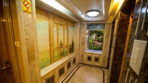 Un couloir doté d’un sol en marbre, d’une fresque murale ornée de colonnes et d’une fenêtre donnant sur un paysage verdoyant.