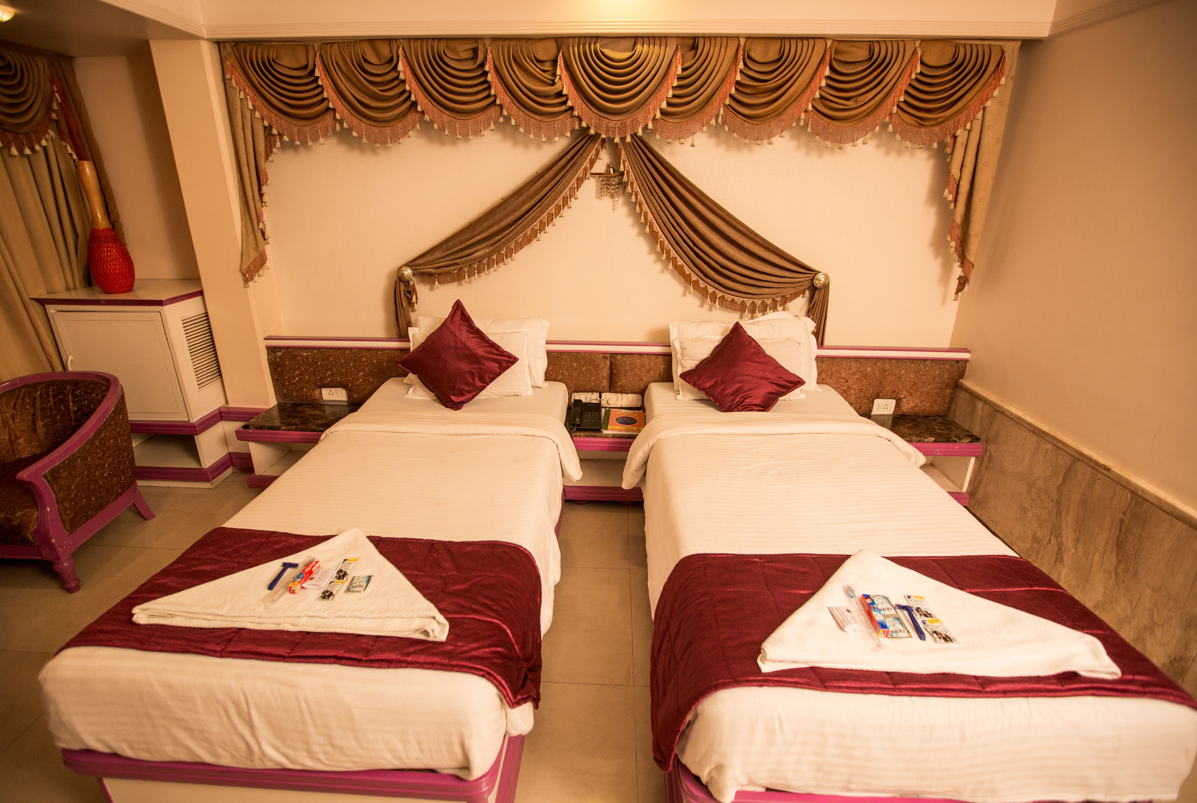 A hotel room with two beds, each with a red bedspread and white pillows. There is a small table between the beds, and a chair to the left.