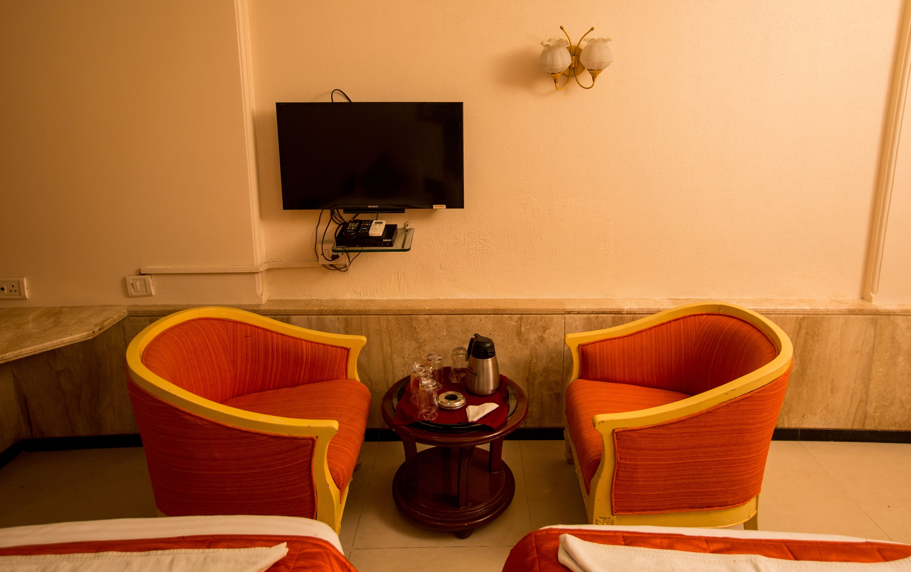 A hotel room with two red upholstered chairs, a small wooden table with a thermos, a flat-screen TV mounted on the wall, and a bed with white bedding.