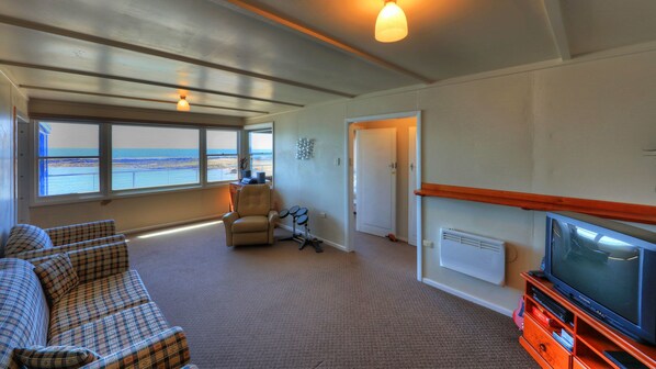 Una sala de estar con un ventanal grande con vista a la playa, un sofá, una silla, un set de batería y un televisor sobre un mueble de madera.