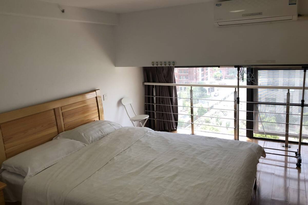 A bedroom with a wooden headboard, a single bed, a small white chair, a window with curtains, and an air conditioning unit.