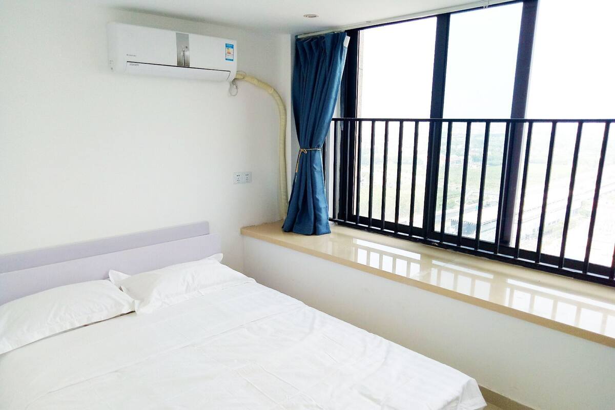 A hotel room with a bed, an air conditioning unit, a window with a view, and a balcony railing.