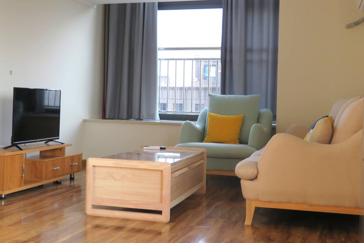 A living room with a wooden coffee table, a sofa, a chair, a TV on a wooden cabinet, and a window with curtains.