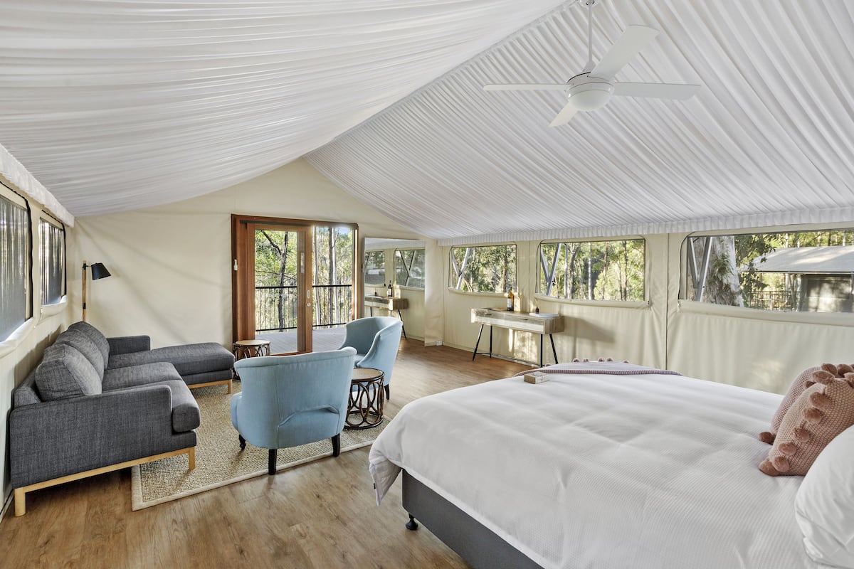 A spacious room with a large bed, a sofa, and a chair. There is a desk and a window with a view of trees.