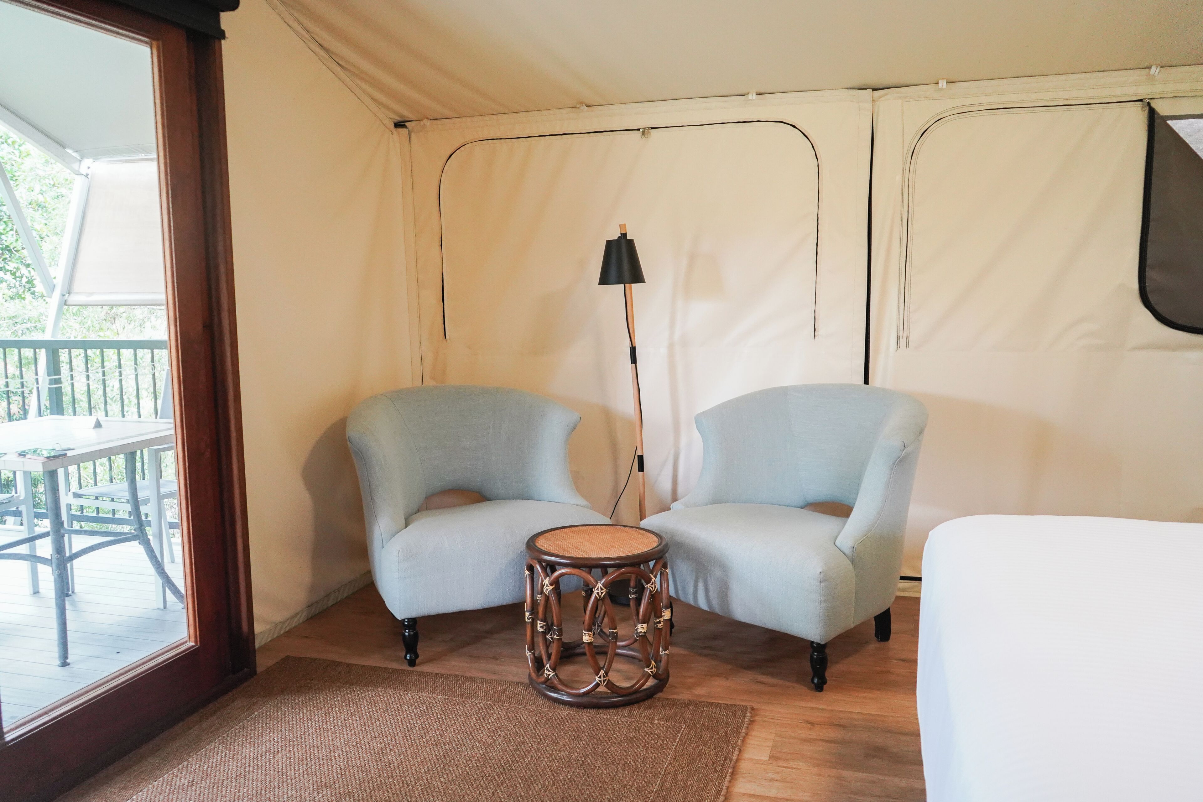 Een tentkamertje met twee lichtblauwe stoelen, een klein rond tafeltje, een vloerlamp en uitzicht op een terras.