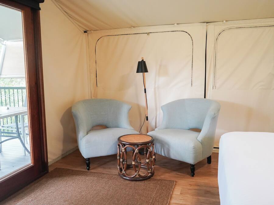 A tented room with two light blue chairs, a small round table, a floor lamp, and a view of an outdoor deck.