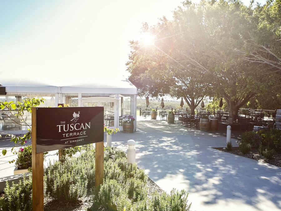 A sign for The Tuscan Terrace at Sirromet, a covered outdoor seating area with tables and chairs, surrounded by greenery and trees.