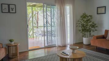 Una sala de estar con una puerta corrediza de vidrio que da a un espacio al aire libre, una planta de bambú en maceta, una mesa redonda de madera y un sofá.