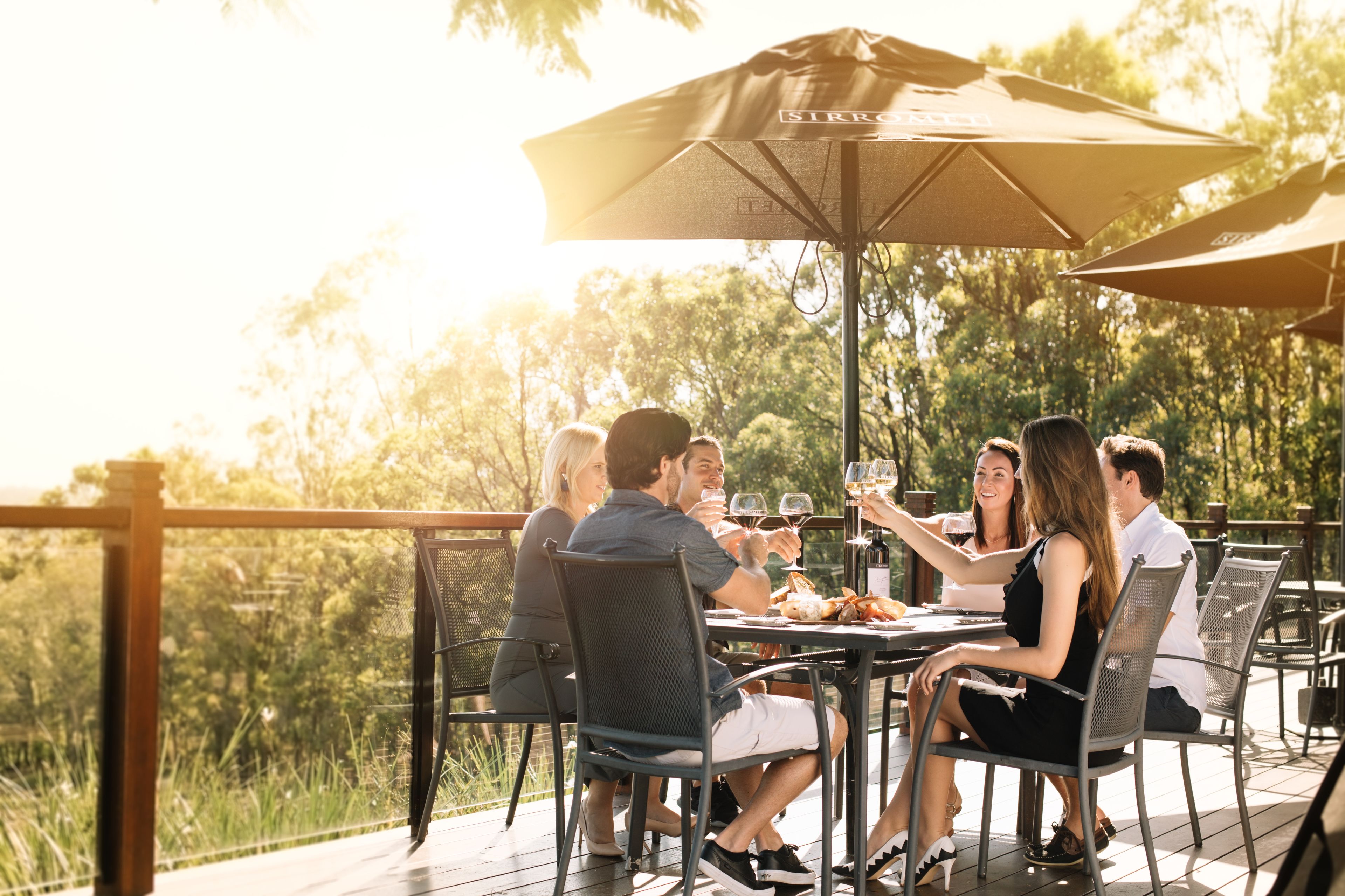 Un grupo de personas cenando en una mesa al aire libre, con sombrillas, rodeado de una terraza de madera y vegetación.
