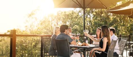 Un grupo de personas cenando en una mesa al aire libre, con sombrillas, rodeado de una terraza de madera y vegetación.