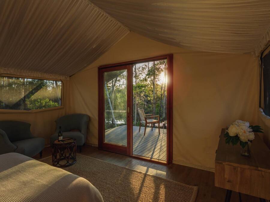 Un dormitorio con cama, dos sillas, una mesita con una botella y ventana con vistas a árboles.