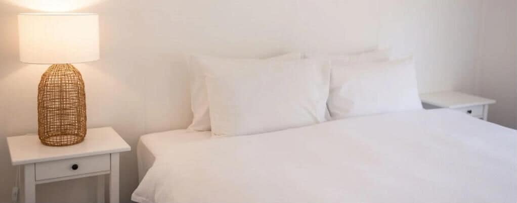 Una cama bien hecha con ropa blanca y almohadas, una lámpara de noche sobre una mesita blanca, y un cajón pequeño.