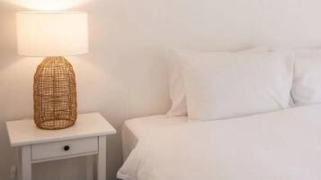 Una cama bien hecha con ropa blanca y almohadas, una lámpara de noche sobre una mesita blanca, y un cajón pequeño.
