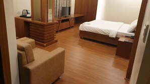 Una habitación de hotel moderna con suelo de madera, cama con ropa blanca, televisor sobre un mueble de madera, sofá y escritorio con hervidor de agua y botellas.