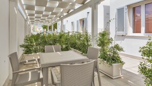 Un patio techado al aire libre con una mesa y sillas, rodeado de plantas en macetas y paredes blancas.