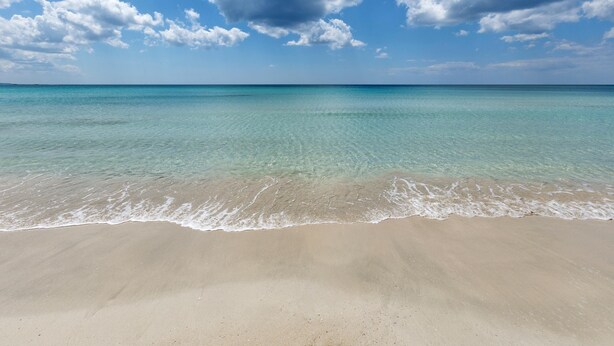 Una playa con agua turquesa cristalina y una orilla arenosa bajo un cielo parcialmente nublado.